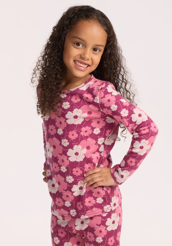 A young girl with long curly hair stands smiling, hands on hips, wearing the Woolx Kids Explorer Base Layer Top in Rosie Posie. She pairs it with matching pants, featuring a pink and white floral pattern against a plain light background. 