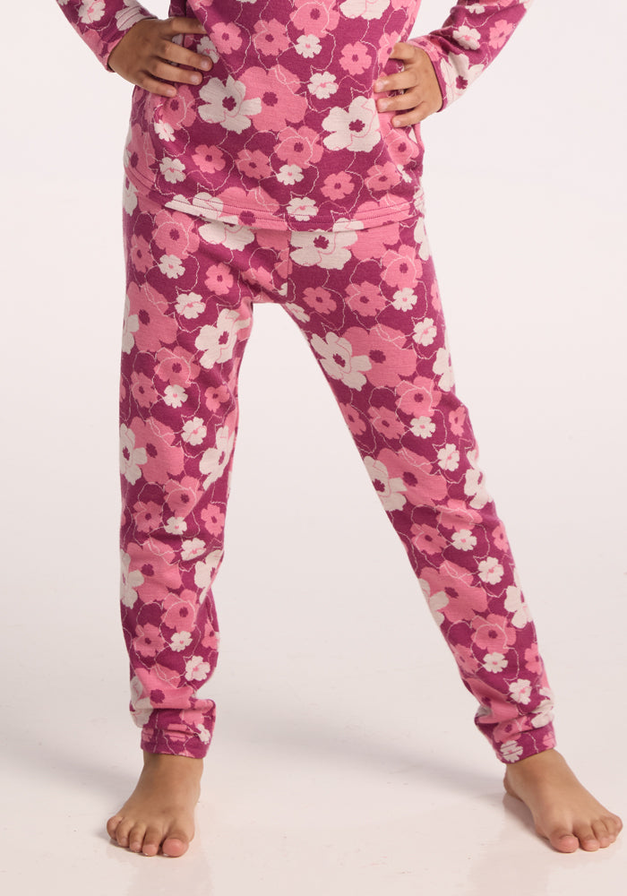 A child stands barefoot, wearing Woolx Kids Explorer Base Layer Bottoms in Rosie Posie under pink and white floral pajamas. The pajamas have long sleeves, and the background is plain and light-colored with the child’s hands on their hips. 