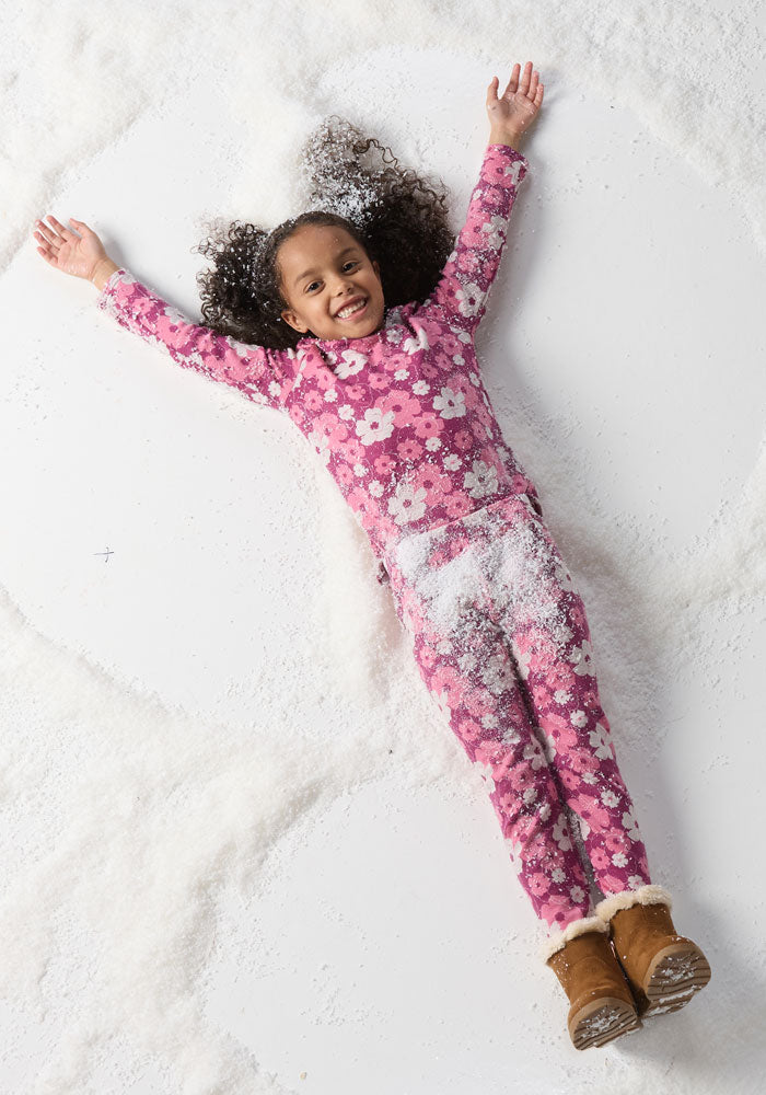 A young child smiles while lying on artificial snow, arms and legs outstretched as if making a snow angel, dressed in the Woolx Kids Explorer Base Layer Top - Rosie Posie.