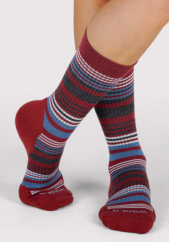 A person wears Woolx Womens Everyday Crew Socks in Rhubarb Stripe, featuring red with blue, white, and black horizontal stripes. The crew socks cover the ankles and lower calves; one foot is slightly raised in front of the other.