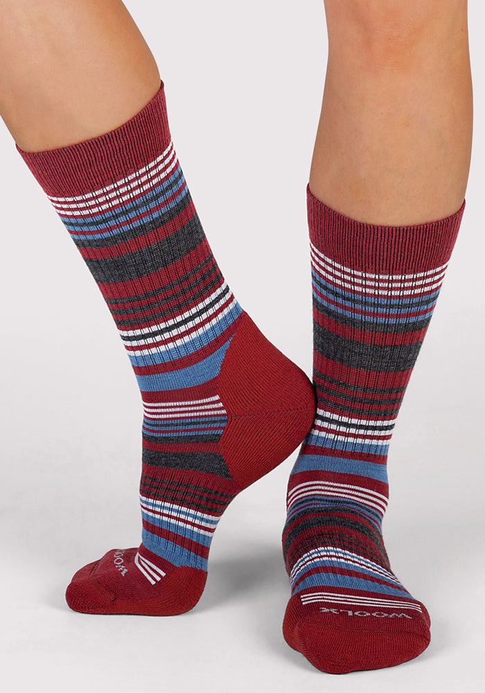 Two feet wearing Woolx Womens Everyday Crew Socks - Rhubarb Stripe, featuring red, blue, white, and black stripes, ribbed texture, and solid red toes and heels. The background is plain and light-colored.
