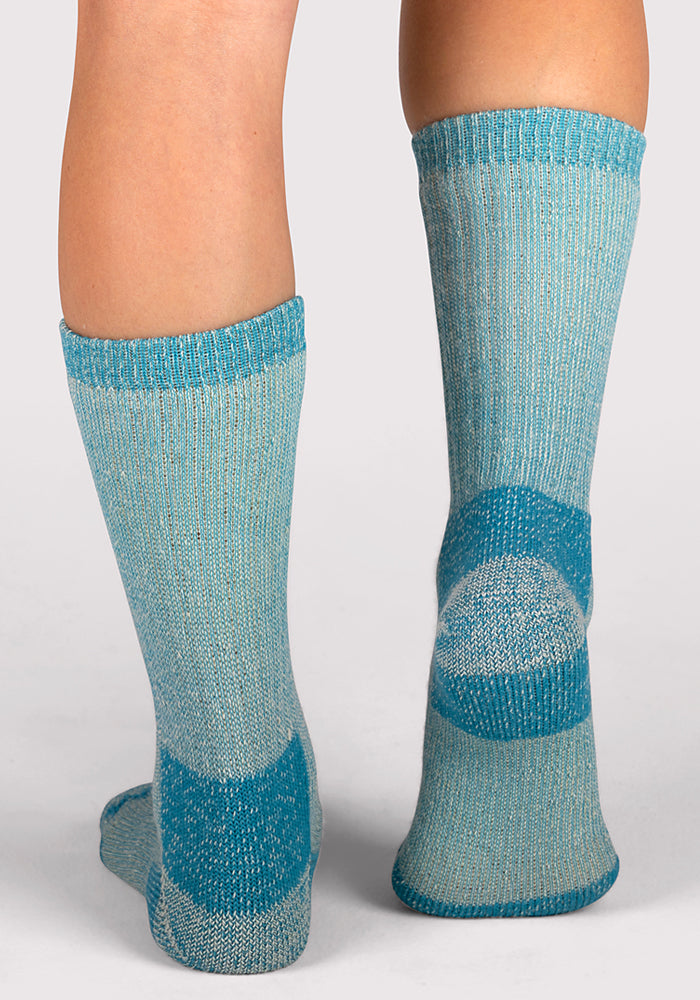 A person wearing Woolx Womens Extreme Crew Socks in Real Teal, featuring darker blue accents on the heel, toe, and top cuff, stands on a light surface, showing the back and side of their calves and feet.