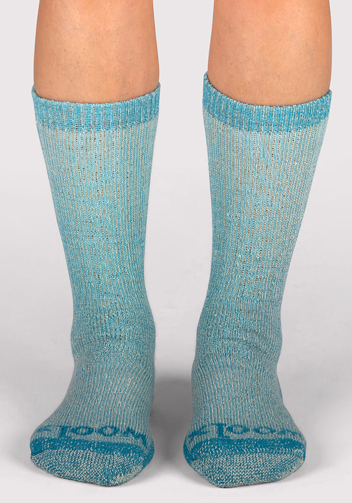 A pair of feet wearing Woolx Womens Extreme Crew Socks in Real Teal, featuring moisture-wicking fabric and darker blue accents at the toes, heels, and cuffs, stands on a plain light-colored surface.