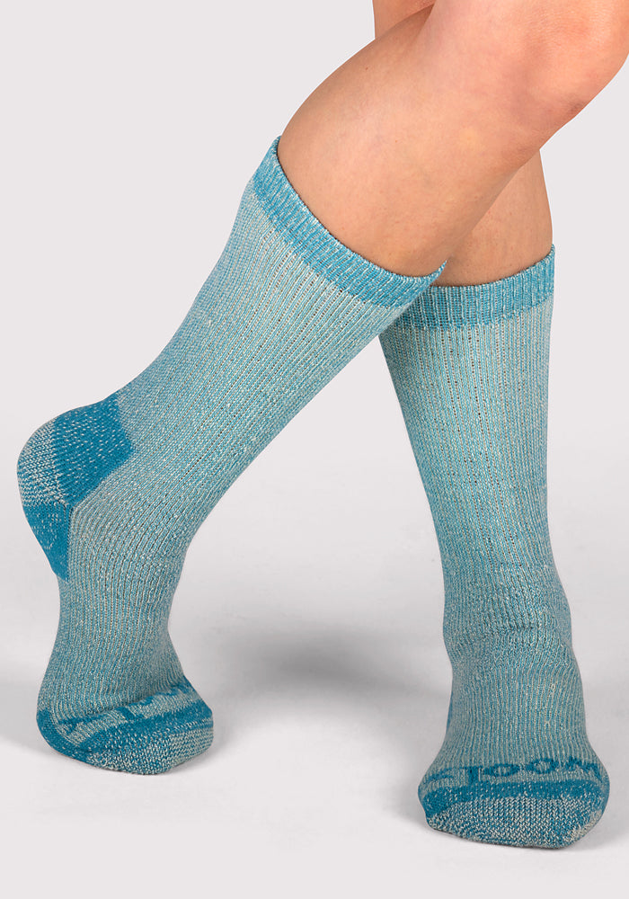 A pair of legs wearing Woolx Womens Extreme Crew Socks in Real Teal, featuring darker blue accents on the toes, heels, and cuffs, stands against a plain light gray background.