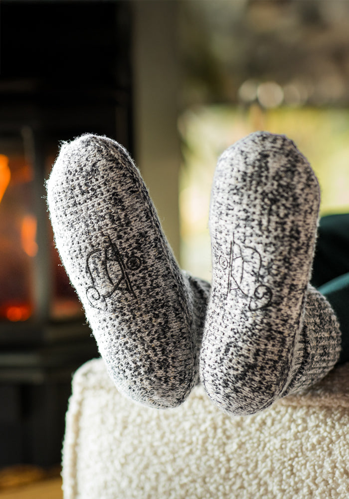 A close-up of feet in Woolx Women’s Gripper Slipper Booties in Speckled Stone, featuring non-slip soles and decorative lettering, resting on an ottoman by a glowing fireplace.