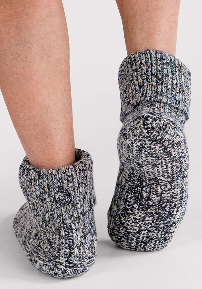 Close-up of feet wearing Woolx Men's Gripper Slipper Bootie in Speckled Stone, featuring thick, moisture-wicking Merino wool and traction soles. Cozy gray and white marled slipper socks are shown against a light background.