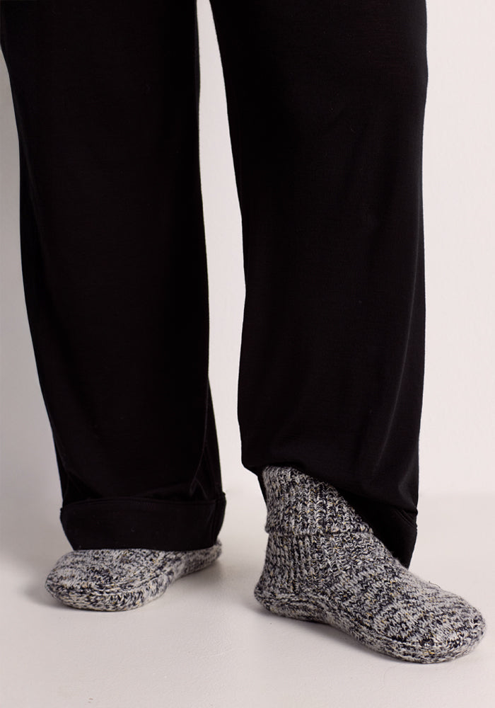 A person wears Woolx Women's Gripper Slipper Booties in Speckled Stone with loose black pants, standing on a light-colored floor; only their lower legs and feet are visible.