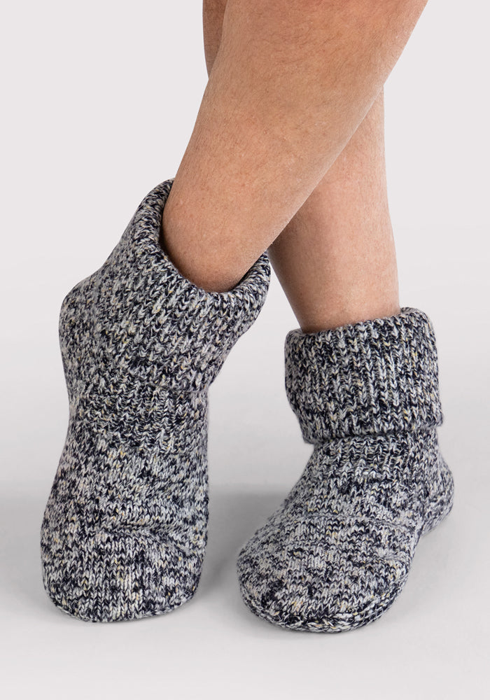 A person wears Woolx Men's Gripper Slipper Bootie in Speckled Stone—cozy, marled gray-and-white Merino socks with cuffs, ankle coverage, textured pattern, and natural odor-resistant comfort—standing on a light background.