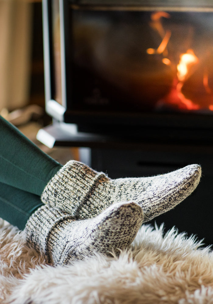 A person wearing Woolx Women's Gripper Slipper Booties in Speckled Stone relaxes with their feet on a plush rug by a cozy fireplace, creating a comfortable and inviting atmosphere.