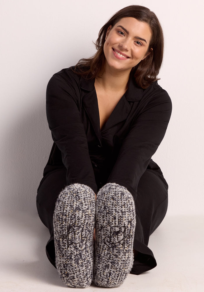 A smiling woman with dark hair, dressed in black, sits on the floor with her knees up, showing the soles of her cozy Woolx Women's Gripper Slipper Booties in Speckled Stone to the camera.