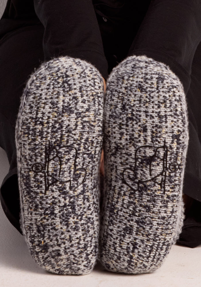 Close-up of the bottoms of two Woolx Women's Gripper Slipper Booties in Speckled Stone with textured soles and abstract cat stitching; person wearing black pants sits with legs bent.