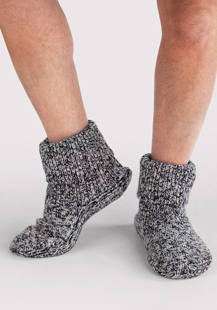 A person wearing Woolx Men's Gripper Slipper Bootie in Speckled Stone stands on a light surface, one foot slightly raised and legs partially visible.