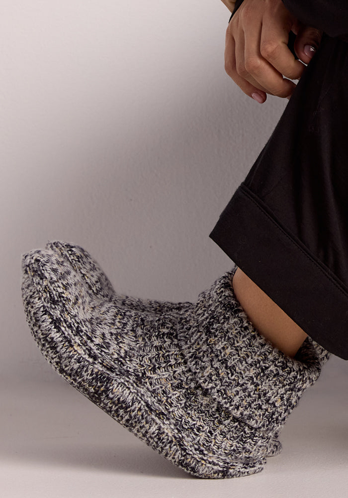 A person wearing Woolx Women's Gripper Slipper Booties in Speckled Stone, featuring chunky knit and cozy sherpa lining, along with a black garment, rests their hand on their knee against a plain light background.