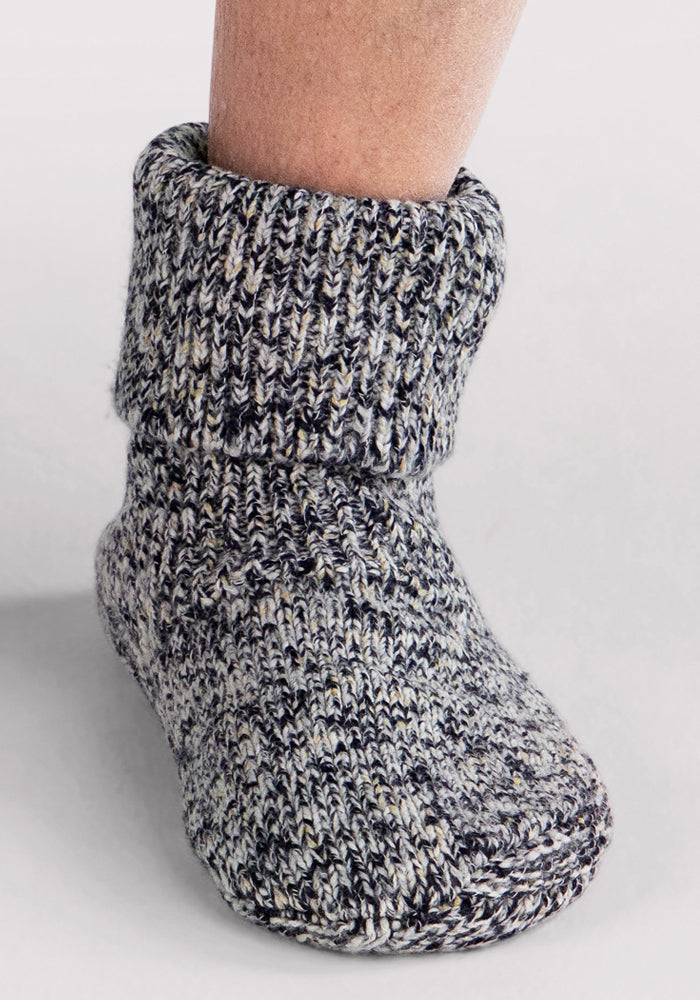A close-up of a person's lower leg and foot wearing the Woolx Men's Gripper Slipper Bootie in Speckled Stone, featuring a thick knit, cozy Merino wool, and a rolled-down top cuff.