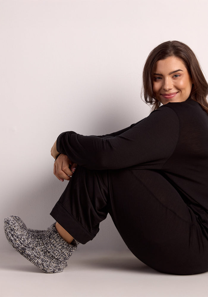 A woman with long brown hair, dressed in black, sits smiling on the floor against a light background, wearing Woolx Women's Gripper Slipper Booties in Speckled Stone with cozy sherpa lining.