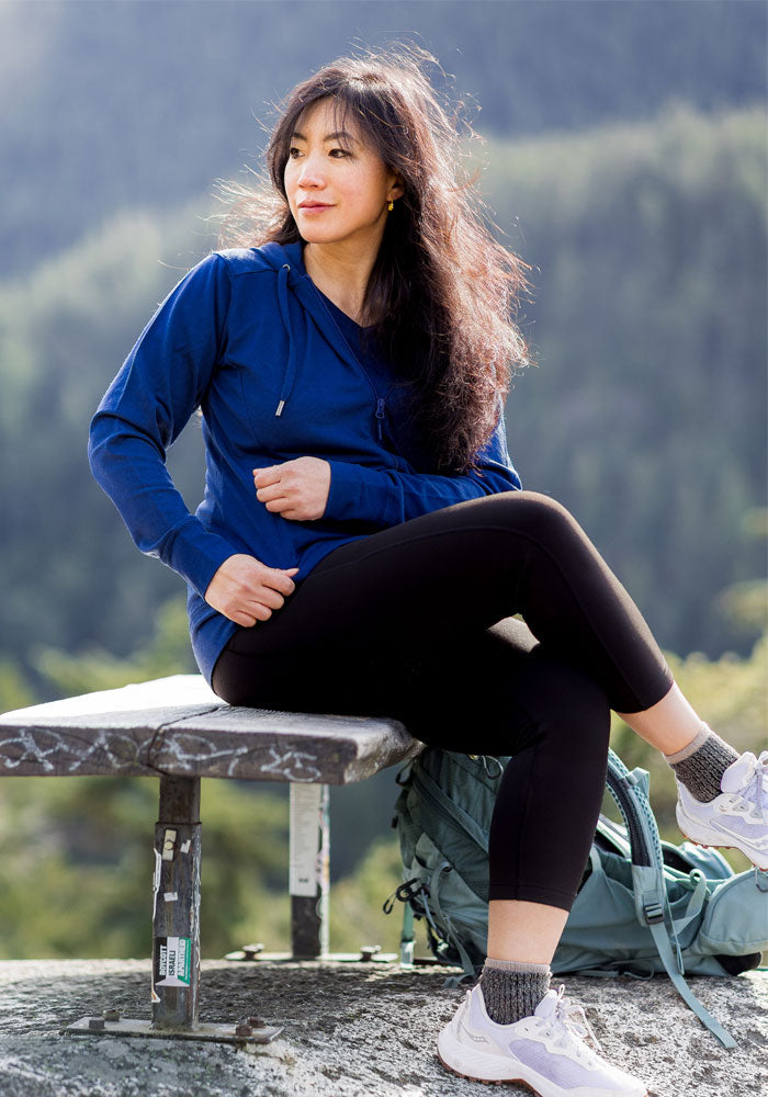 A woman with long dark hair wears the Woolx Ryann Hoodie in Estate Blue, paired with black leggings and sneakers. She sits on a bench outdoors beside a green backpack, forested mountains behind her—a true travel essential scene.