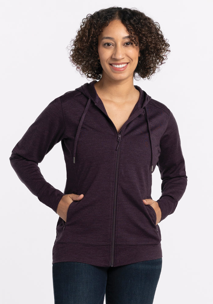 A person with curly hair smiles in a deep plum Ryann Hoodie by Woolx, crafted from lightweight merino wool, paired with dark jeans. With hands in pockets against a white background, they embody a travel-ready design perfect for any adventure.