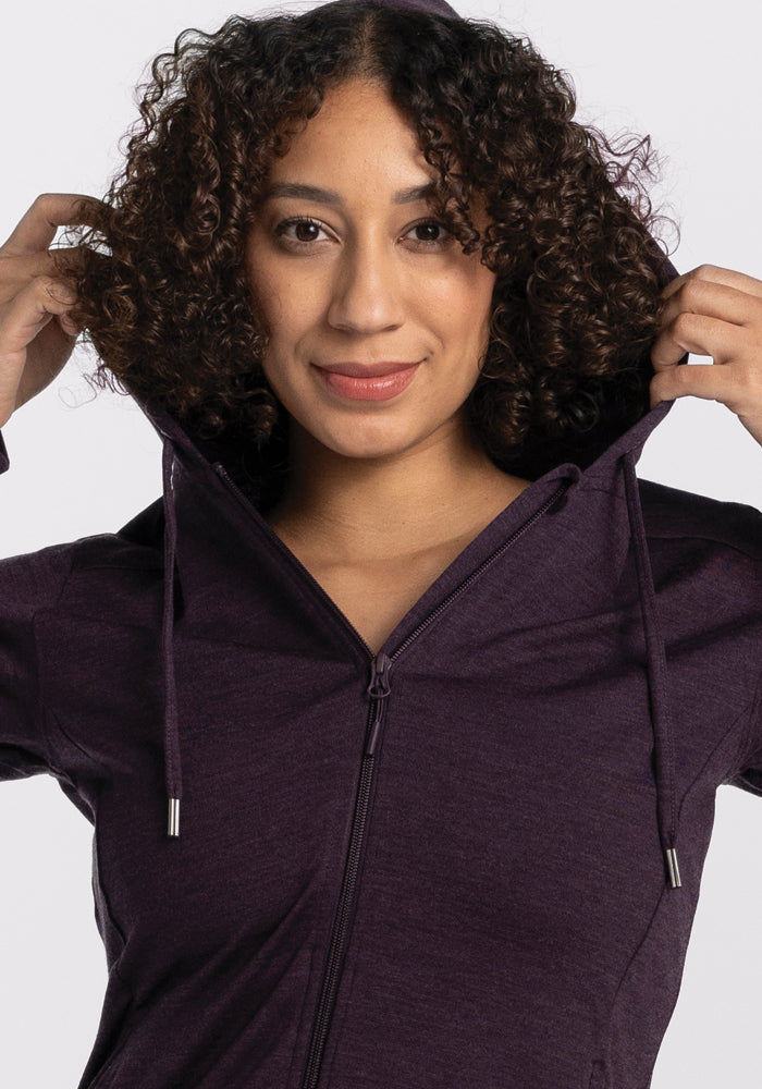 A person with curly hair adjusts the hood of a Woolx Ryann Hoodie in Deep Plum, smiling slightly. The travel-ready design pops against the plain, light-colored background.