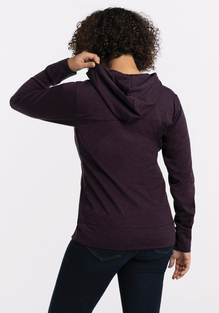 A curly-haired individual, dressed in a Woolx Ryann Hoodie - Deep Plum and dark jeans, faces away from the camera. They hold the hood with their right hand against a plain white background.