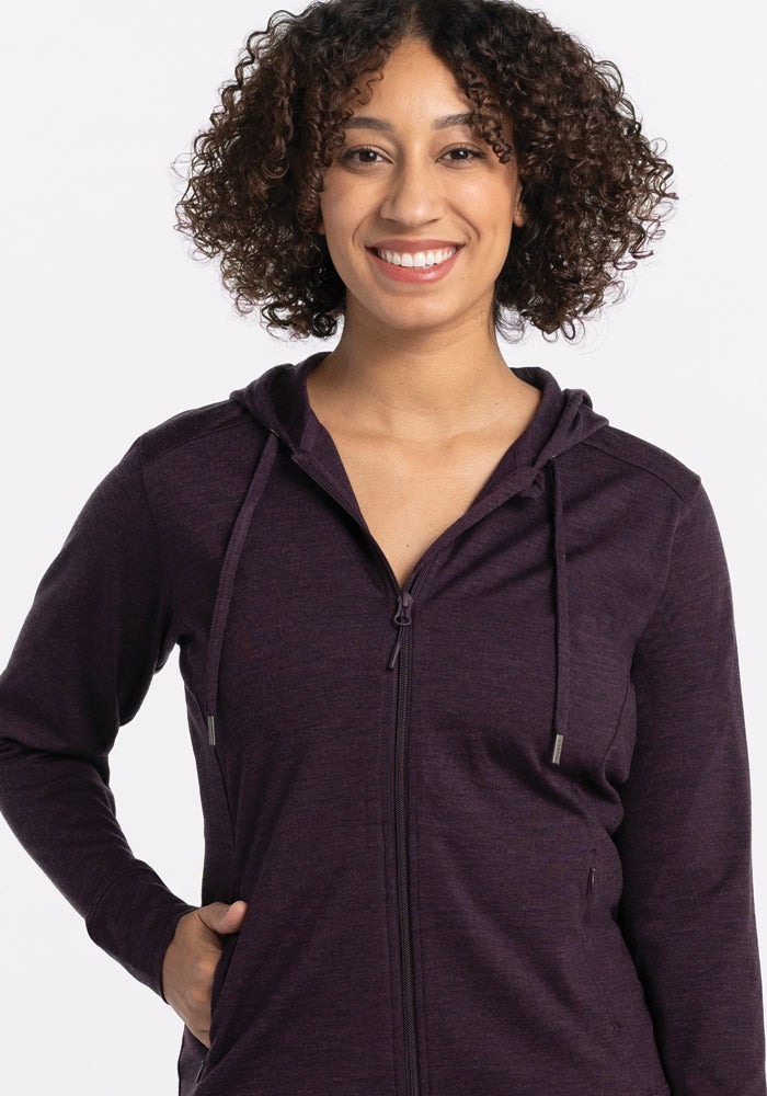 A curly-haired person smiles at the camera, wearing the lightweight Woolx Ryann Hoodie, featuring a deep plum color and zip-up design with one hand in a pocket. The background is plain white.