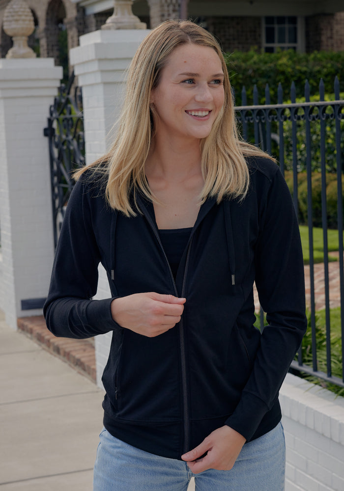 A woman with long blonde hair, wearing the Woolx Ryann Hoodie in Cream Heather and light blue jeans, stands outside smiling by a white brick wall and black iron fence—perfect for a travel essential photo.