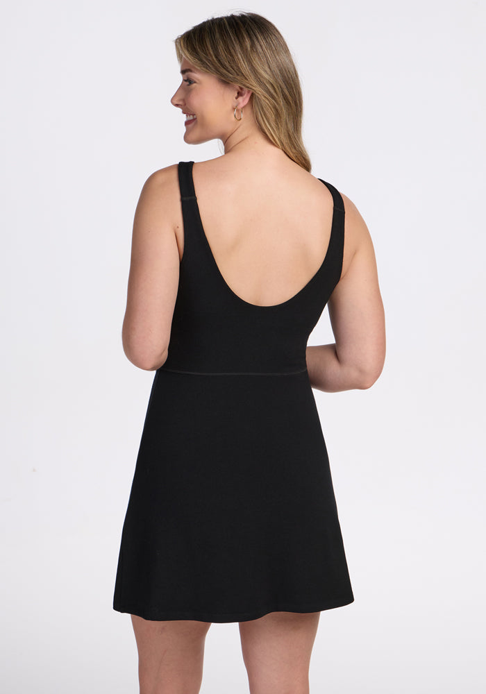 A woman with long, light brown hair stands facing away, smiling to her left. She wears the Woolx Adrienne Active Dress - Black, a sleeveless athletic dress with built-in shorts, set against a plain white background.
