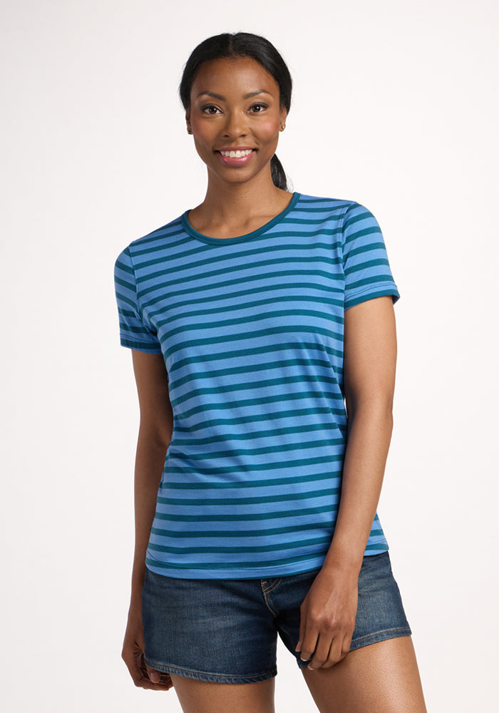 A woman stands against a plain background, smiling. She wears the Woolx Liza Crew Neck Tee - Coastal Stripe and denim shorts. Her hair is pulled back in a low ponytail. 