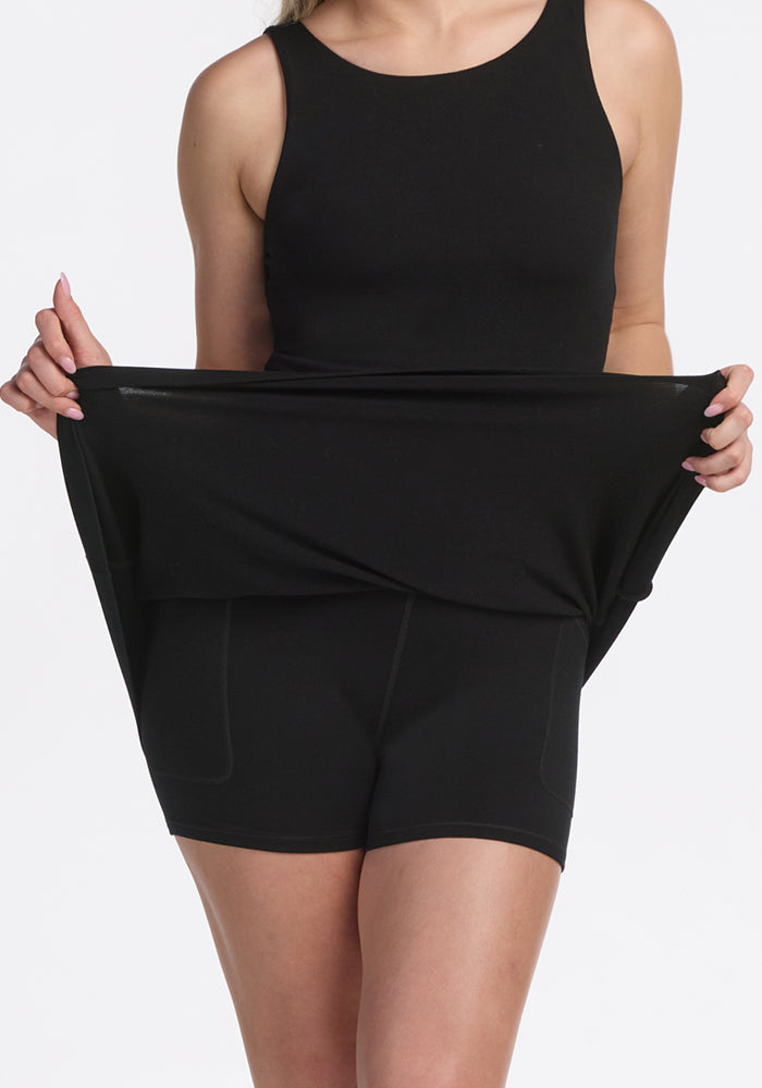 A woman lifts the hem of the black Woolx Adrienne Active Dress to show its built-in shorts, set against a plain white background.