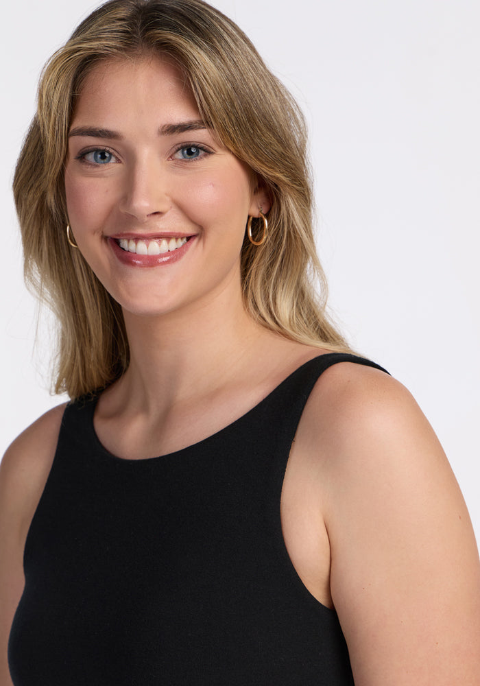 A woman with long, light brown hair and blue eyes smiles at the camera, wearing the Woolx Adrienne Active Dress - Black, a sleeveless athletic dress with built-in shorts and gold hoop earrings, set against a plain white background.