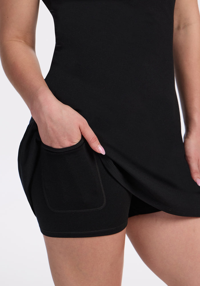 A person wearing the Woolx Adrienne Active Dress - Black lifts the hem to reveal black shorts with a pocket, where their hand rests, against a plain light background.