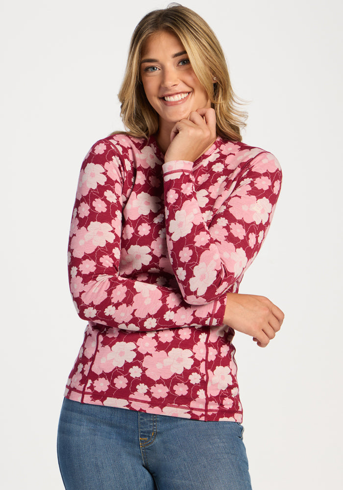 Smiling blonde woman wears Woolx Maddie Baselayer Top in Floral Crush and blue jeans, hand near her face. 