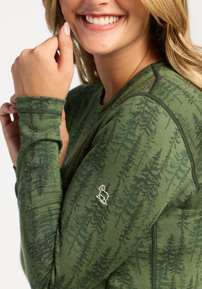 Woman smiling, wearing the Woolx Maddie Baselayer Top - Forest Friends, featuring a green forest tree pattern.