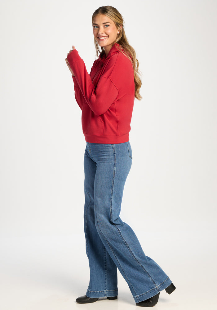 Woman in Woolx Lizzy Hoodie - Maraschino Cherry and flared blue jeans stands sideways on a white background.