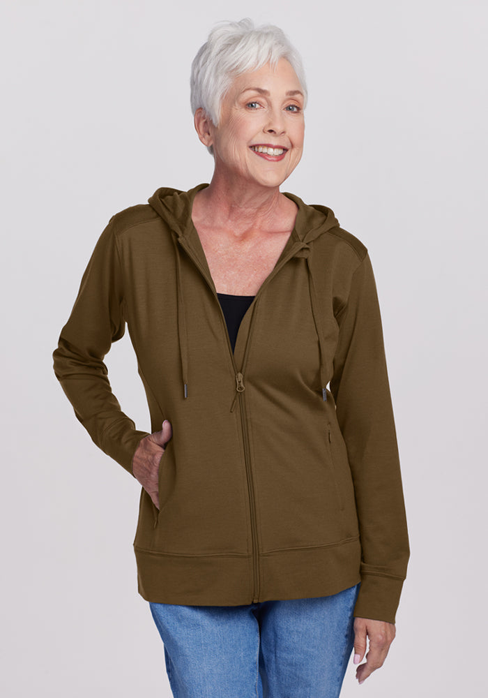 Smiling older woman wears Woolx Ryann Hoodie in Olive Leaf and blue jeans, a perfect travel essential, on plain backdrop. | Kathy is 5’9”, wearing a size S