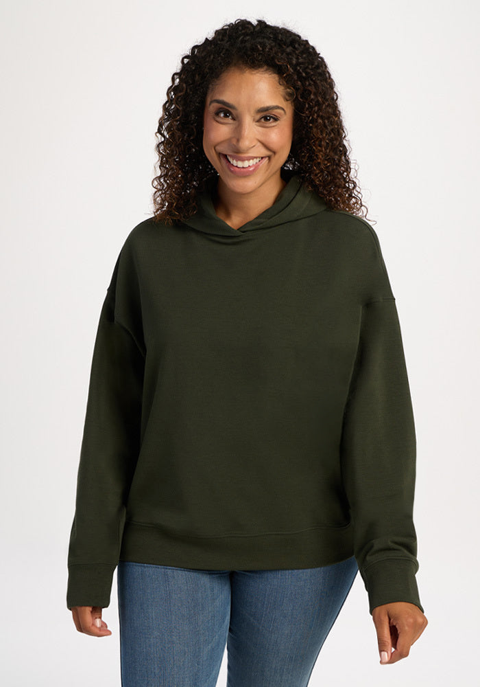 A smiling woman wears an olive green Woolx Genevieve Hoodie - Duffel Bag and blue jeans. 