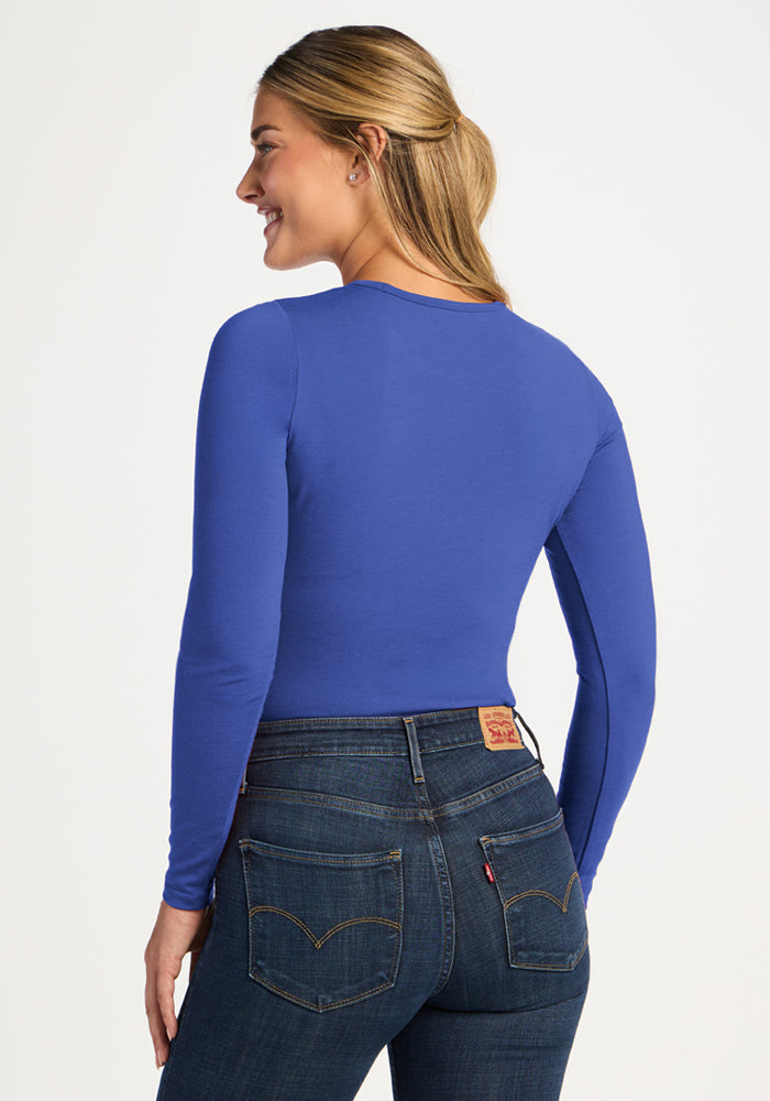 Woman in a Woolx Margot Bodysuit - Royal Blue and dark jeans, facing away and smiling, with light brown hair.
