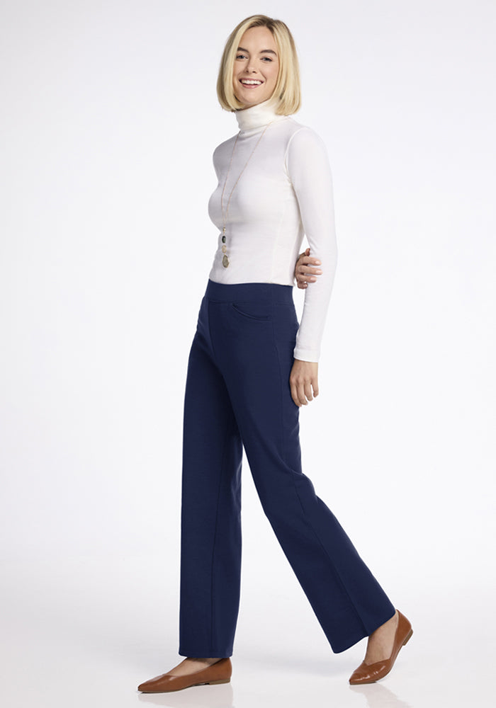 Petite woman in Woolx Ellie Wide Leg Pants Petite - Ocean Cavern, white turtleneck, and brown flats smiles on white.