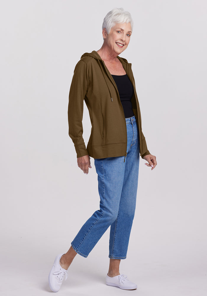 Smiling older woman in a Woolx Ryann Hoodie - Olive Leaf, black top, blue jeans, and white sneakers against plain background.