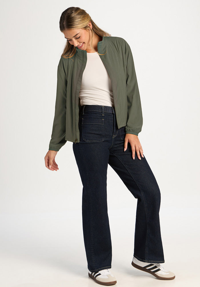 A woman wears a sage olive Cooper Windbreaker over a white shirt and dark jeans, styled in an all-season woven fabric.