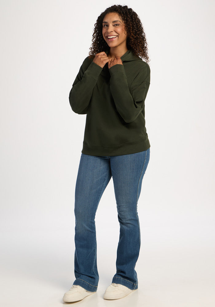 A smiling woman wearing a dark green Woolx Genevieve Hoodie - Duffel Bag, blue jeans, and white sneakers.