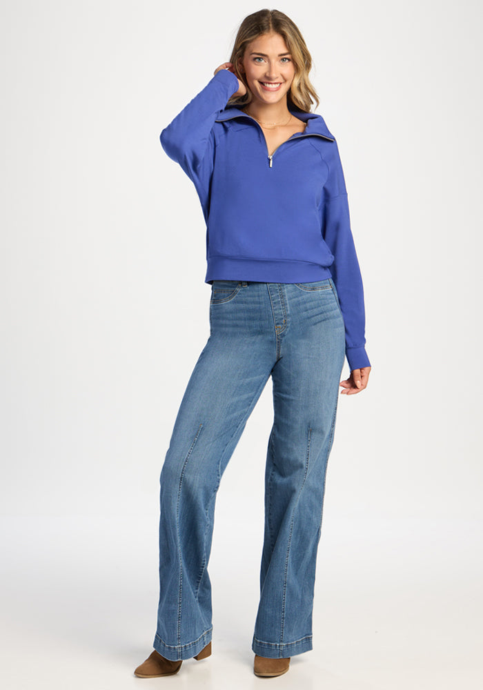 A smiling woman wears a royal blue Woolx Tori 1/4 Zip and blue wide-leg jeans.