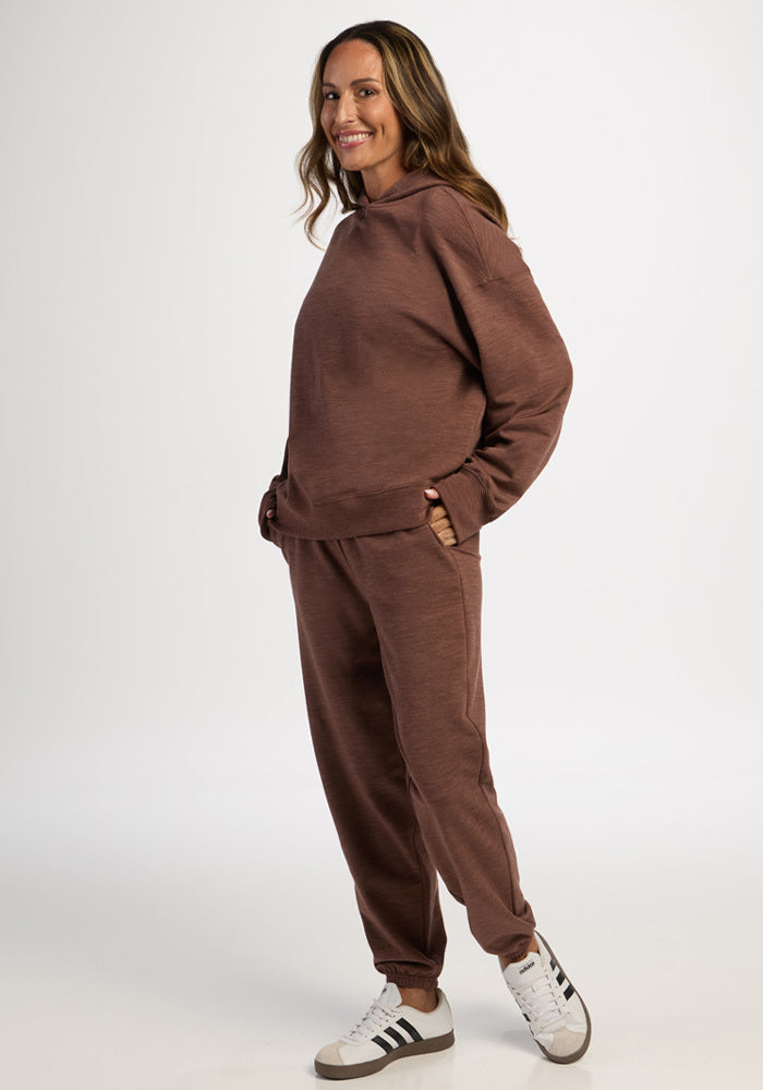 A woman wearing a brown moisture-wicking Genevieve Hoodie - Sable Heather and matching joggers, hands in her pockets.