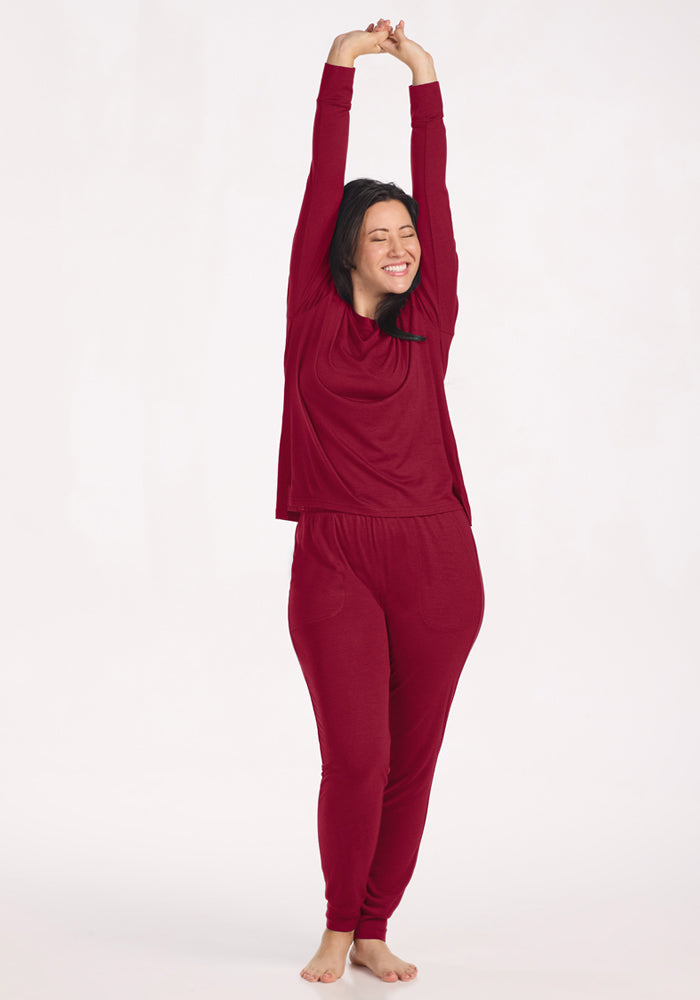 A smiling woman in a long-sleeved shirt and beet red Mary Kate Lounge Pants stretches her arms above her head.