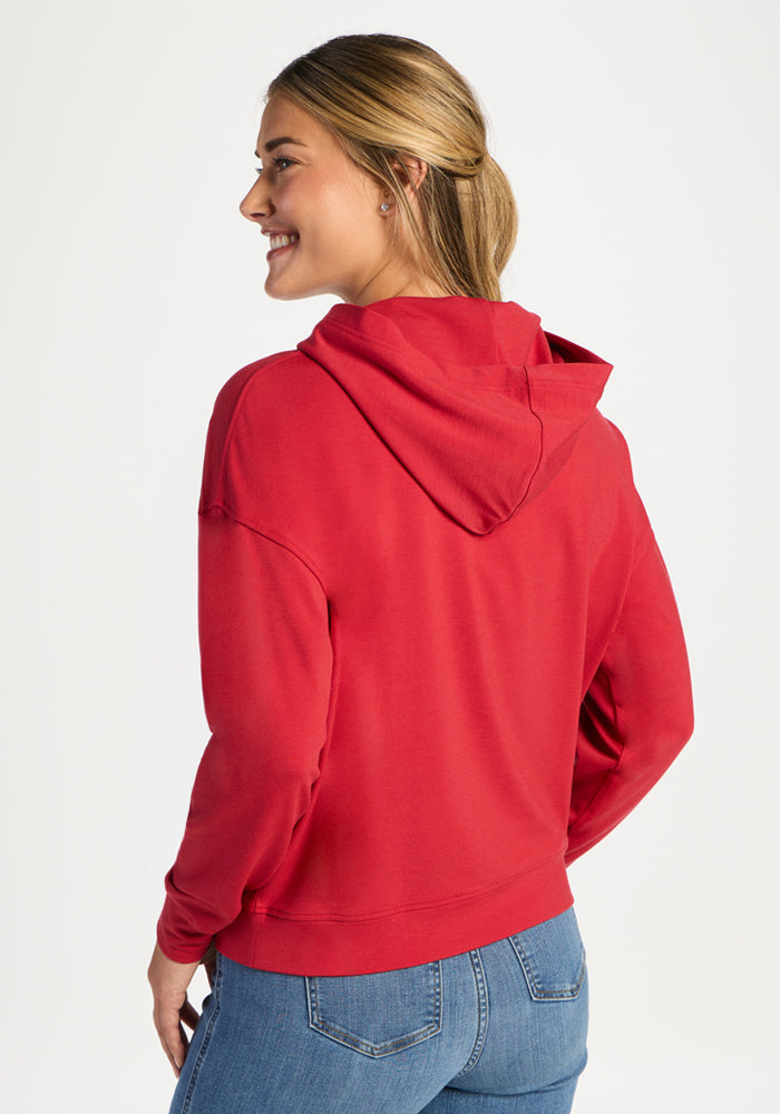 Woman in a Woolx Lizzy Hoodie - Maraschino Cherry and blue jeans smiles, looking over her shoulder against white.