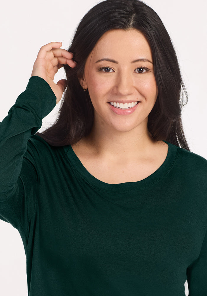 A smiling woman wears a long-sleeved dark green Woolx Ashlie Lounge Top in Deep Pine with a round neckline.