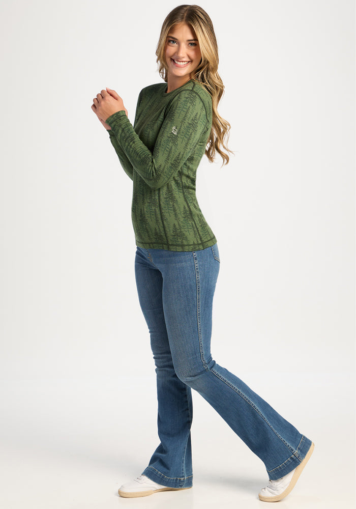 Smiling woman wears Woolx Maddie Baselayer Top - Forest Friends and blue jeans, posing with raised arms on white background.