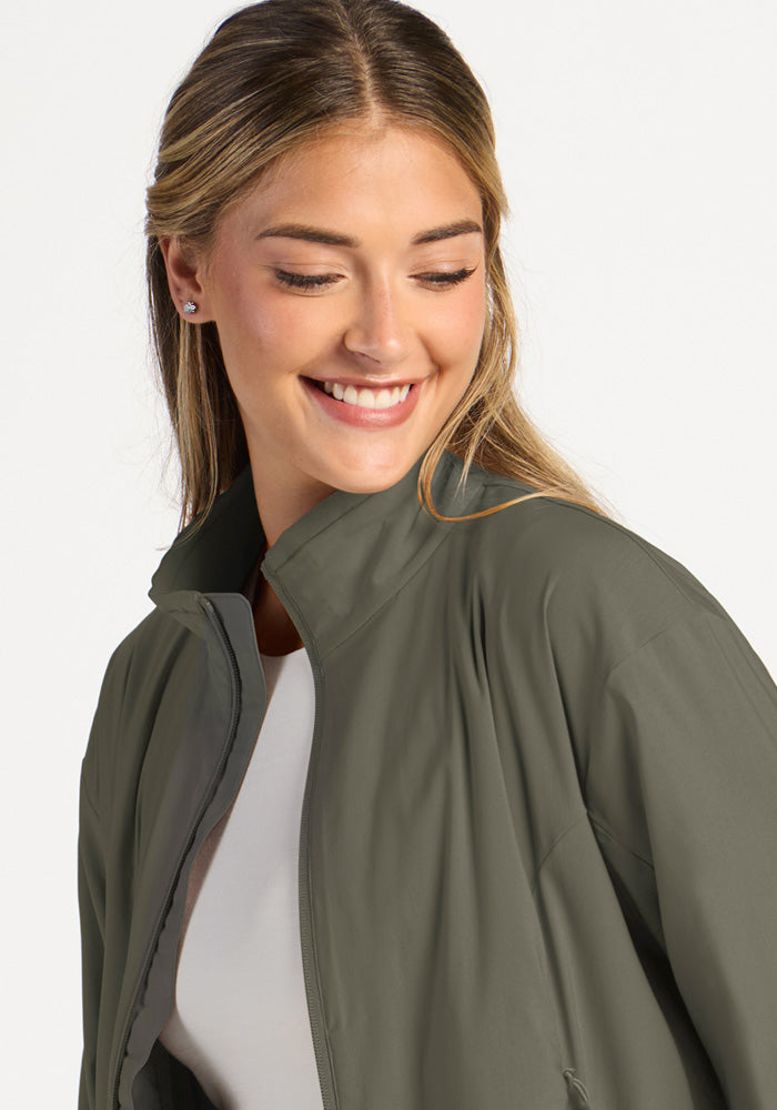 A smiling woman wears a sage olive Woolx Cooper Windbreaker with a high collar over a white shirt.