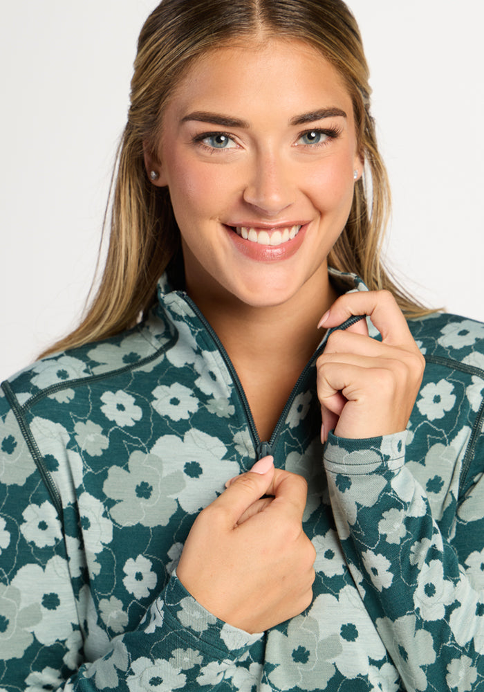 Smiling woman with long hair wears Woolx Emi 1/4 Zip - Coastal Wildflower, holding the zipper near her chin.