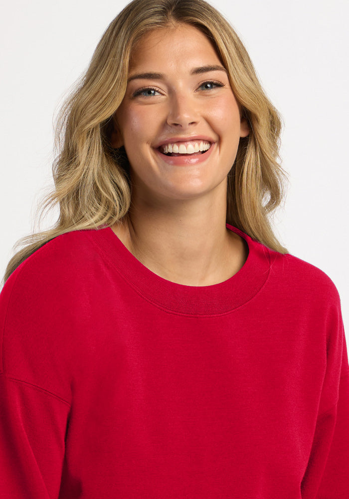 A woman smiles wearing a bright red Molly Crewneck - Maraschino Cherry, a temperature-regulating merino wool sweatshirt.