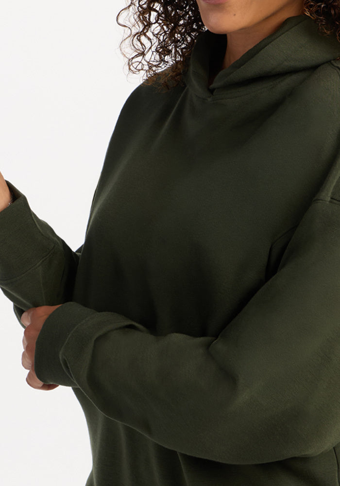 A person wearing a dark green Woolx Genevieve Hoodie - Duffel Bag, a temperature-regulating merino wool hooded sweatshirt.